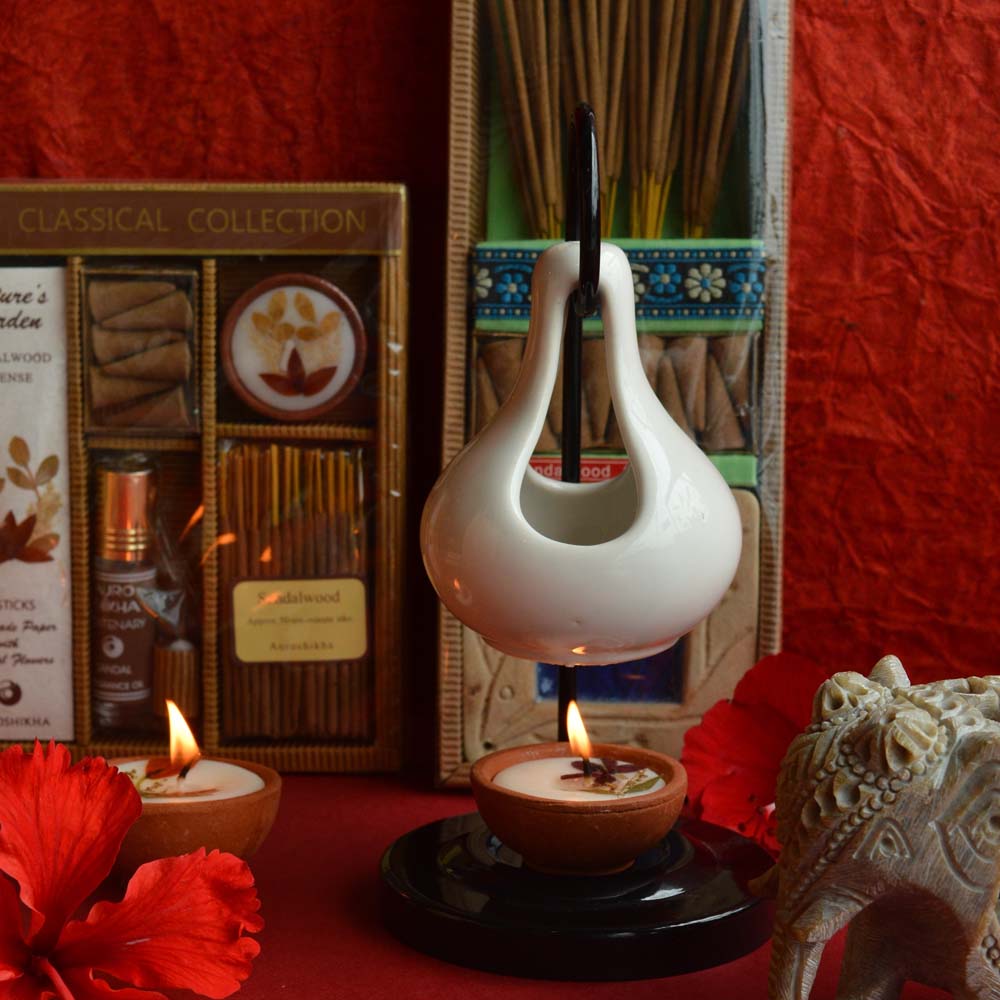 Cera White Ceramic Oil Diffuser with a lit tealight, surrounded by incense sticks, candles, and a carved elephant figurine on a vibrant red background.