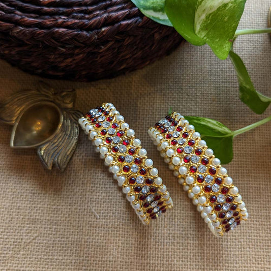 Gold bangles with ruby red stones and pearl detailing, placed on a rustic background with green leaves and brass decor.