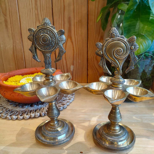 Set of traditional brass Shank Chakra lamps with intricate designs, displayed on a wooden surface alongside a decorative pot with marigold flowers.