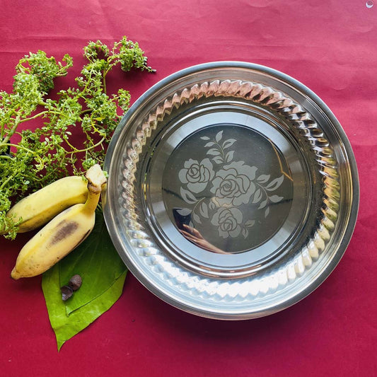 Stainless steel puja plate with floral engraving, placed on a red background, surrounded by green leaves and bananas.