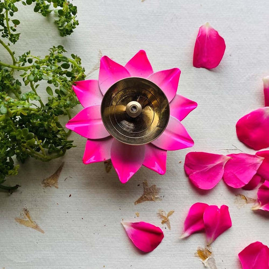 Handcrafted pink lotus diya with a brass center, surrounded by greenery and rose petals on a white textured surface.