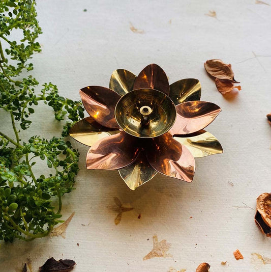 Handcrafted copper-brass lotus diya (oil lamp) with a dual-tone finish, surrounded by greenery and dried petals on a white textured surface.