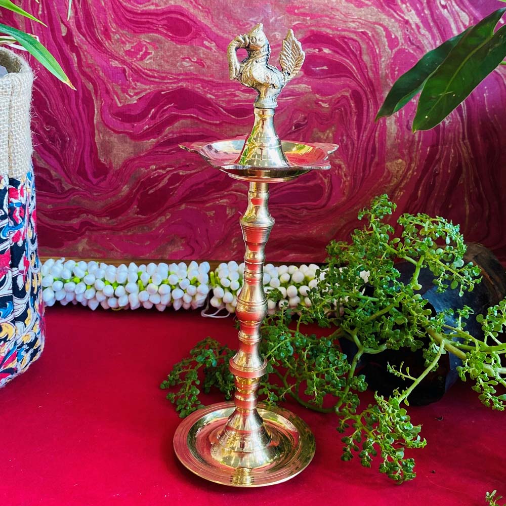 Traditional brass kuthu vilakku (oil lamp) with a peacock design, surrounded by plants and vibrant decor on a red surface.