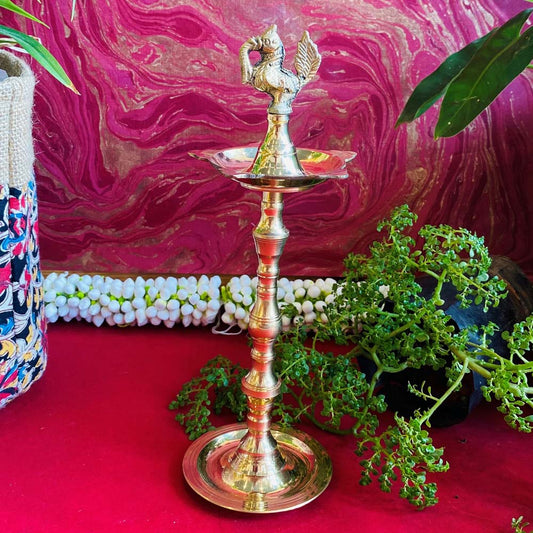 Traditional brass kuthu vilakku (oil lamp) with a peacock design, surrounded by plants and vibrant decor on a red surface.