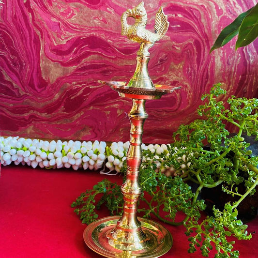 Traditional brass kuthu vilakku (oil lamp) with a peacock design, surrounded by plants and vibrant decor on a red surface.