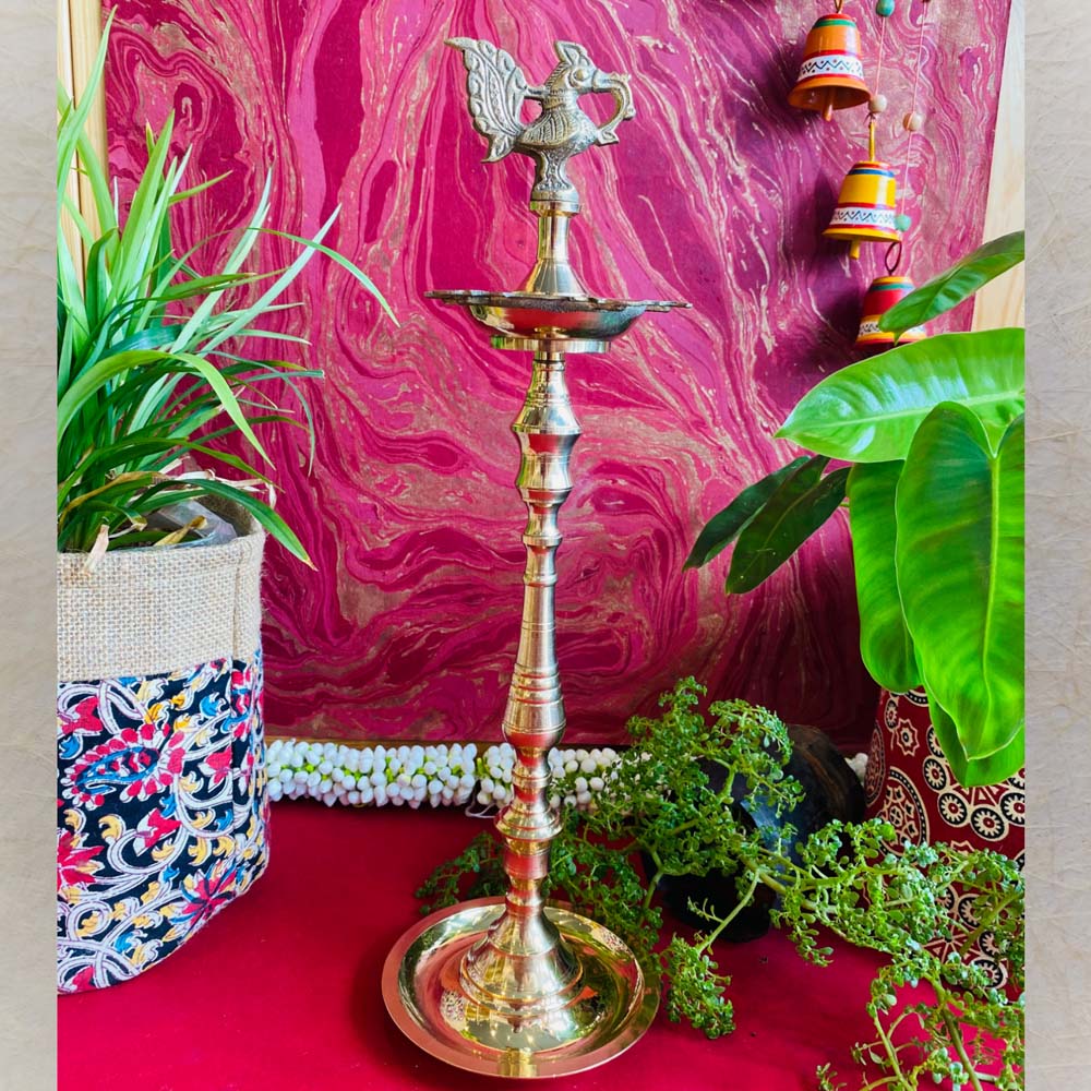 Traditional brass kuthu vilakku (oil lamp) with a peacock design, surrounded by plants and vibrant decor on a red surface.