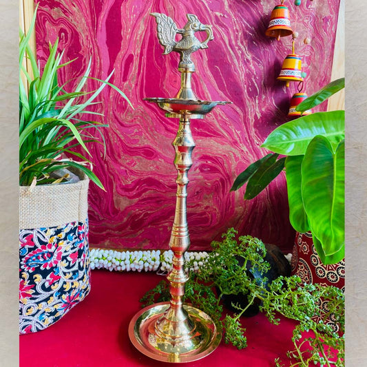 Traditional brass kuthu vilakku (oil lamp) with a peacock design, surrounded by plants and vibrant decor on a red surface.