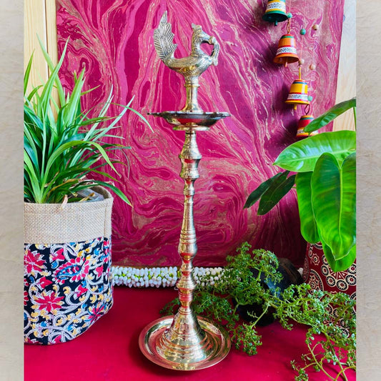 Traditional brass kuthu vilakku (oil lamp) with a peacock design, surrounded by plants and vibrant decor on a red surface.