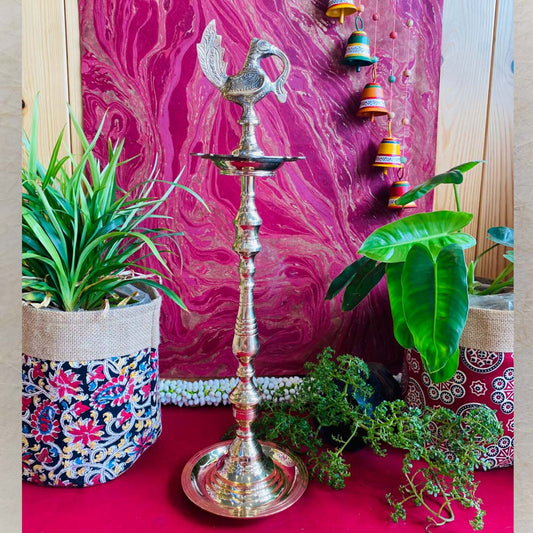 Traditional brass kuthu vilakku (oil lamp) with a peacock design, surrounded by plants and vibrant decor on a red surface.