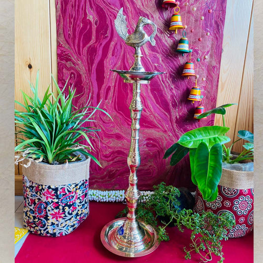 Traditional brass kuthu vilakku (oil lamp) with a peacock design, surrounded by plants and vibrant decor on a red surface.
