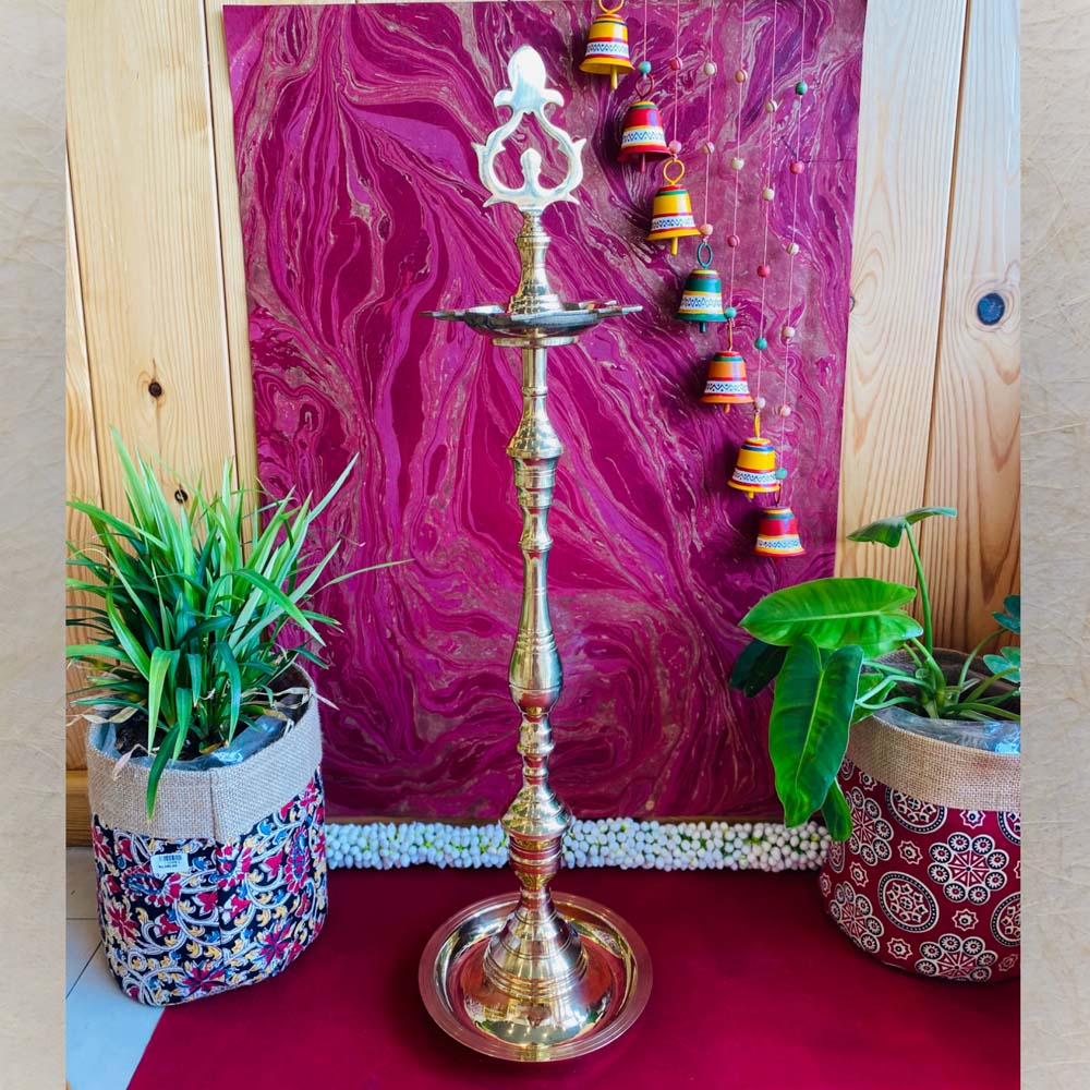 Traditional brass kuthu vilakku (oil lamp) with an intricate top design, surrounded by plants and vibrant decor on a red surface.