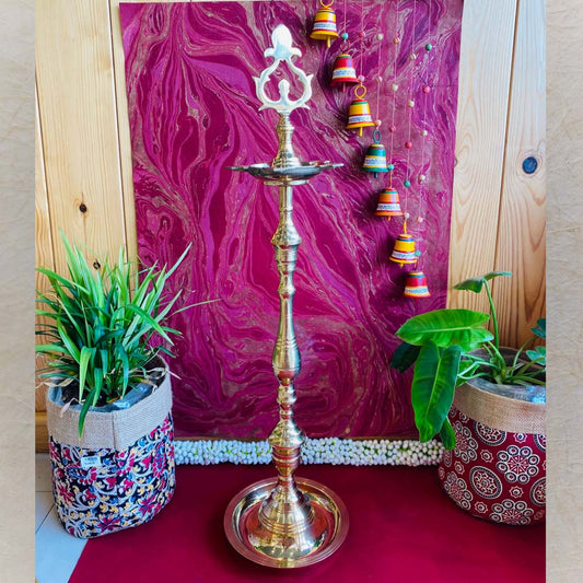 Traditional brass kuthu vilakku (oil lamp) with an intricate top design, surrounded by plants and vibrant decor on a red surface.