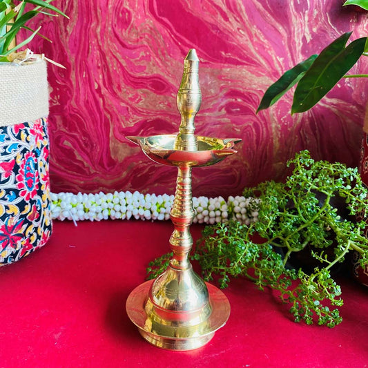 Handcrafted brass Kerala lamp (Nilavilakku) with a polished finish, placed on a vibrant red surface with plants and colorful decor in the background.