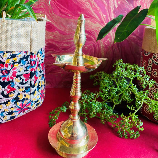 Handcrafted brass Kerala lamp (Nilavilakku) with a polished finish, placed on a vibrant red surface with plants and colorful decor in the background.