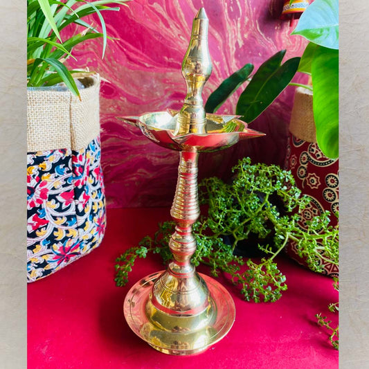Handcrafted brass Kerala lamp (Nilavilakku) with a polished finish, placed on a vibrant red surface with plants and colorful decor in the background.