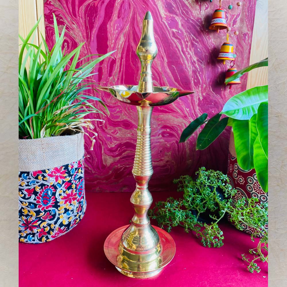 Handcrafted brass Kerala lamp (Nilavilakku) with a polished finish, placed on a vibrant red surface with plants and colorful decor in the background.