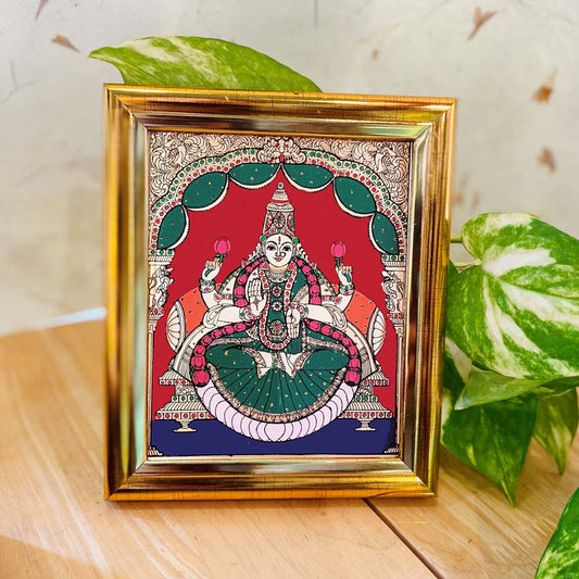 Tanjore-style artwork of Goddess Lakshmi seated on a lotus, holding lotus flowers, set against a red and green backdrop and framed in a gold-colored frame.