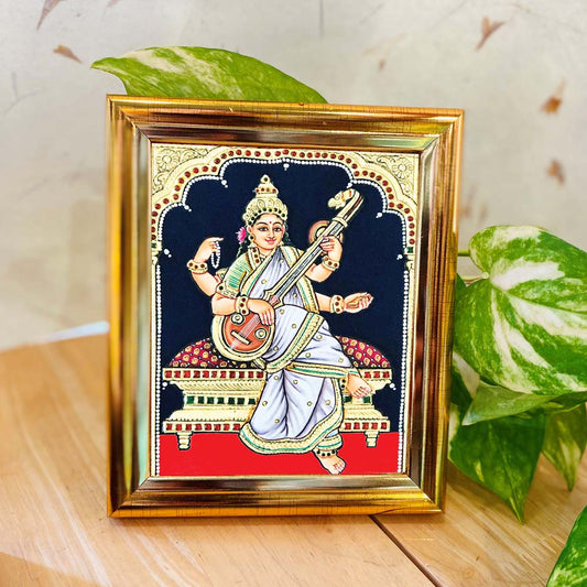Tanjore-style artwork of Goddess Saraswathi seated with a veena, adorned with intricate details, set against a black backdrop, and enclosed in a golden frame.