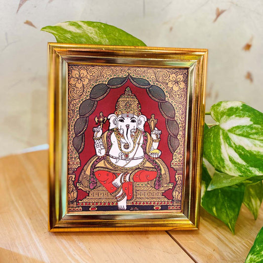 Tanjore-style artwork of Lord Ganesha seated gracefully with vibrant gold embellishments, set against a maroon backdrop and enclosed in a golden frame.