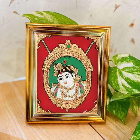 Tanjore-style artwork of Lord Krishna depicted in a pendant-like frame design, set against a red and green background and enclosed in a gold-colored frame.