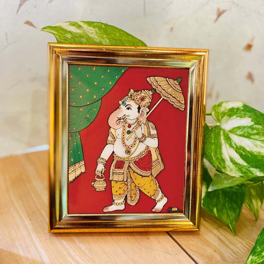 Tanjore-style artwork of Lord Ganesha holding an umbrella and lamp, set against a red and green backdrop in a gold-colored frame.