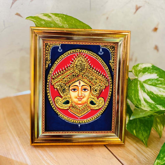 Tanjore-style artwork of Goddess Durga's face adorned with a golden crown and intricate details, set against a red and blue backdrop in a gold-colored frame.