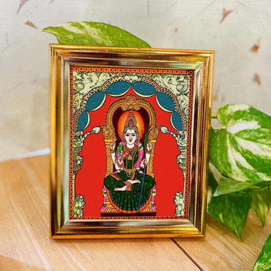 Tanjore-style artwork of Goddess Kamakshi seated in splendor, set against a red backdrop with a decorative gold frame.