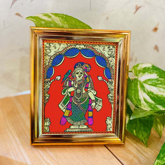 Tanjore-style framed artwork featuring Goddess Andal in a vibrant red backdrop, adorned with intricate detailing and enclosed in a golden frame, placed on a wooden surface.