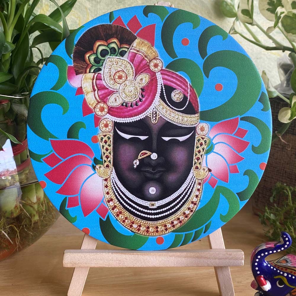 Decorative plate featuring the face of Shreenathji adorned with intricate jewelry and vibrant floral patterns, set on a blue background and displayed on a wooden easel.