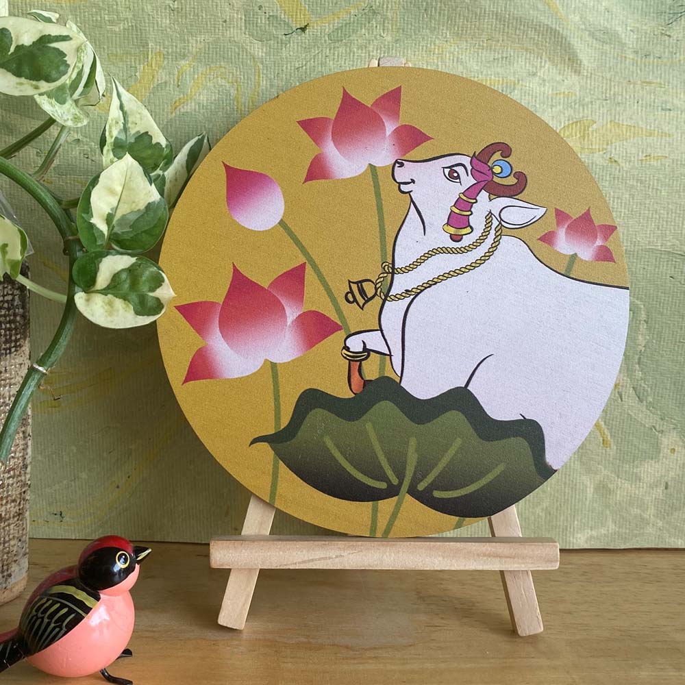 Hand-painted decorative plate featuring a white cow adorned with traditional jewelry, surrounded by pink lotus flowers, set against a yellow background and displayed on a wooden easel.