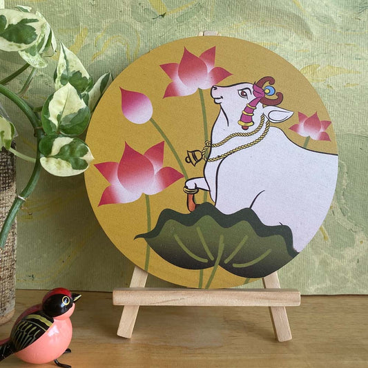 Hand-painted decorative plate featuring a white cow adorned with traditional jewelry, surrounded by pink lotus flowers, set against a yellow background and displayed on a wooden easel.