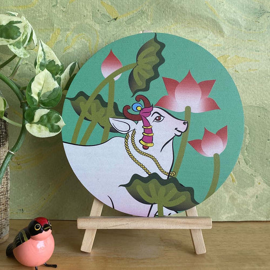 Hand-painted decorative plate featuring a white cow surrounded by pink lotus flowers and lush green leaves, set against a green background and displayed on a wooden easel.