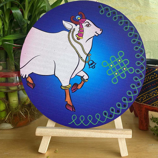 Hand-painted decorative plate with a white cow adorned with traditional jewelry, set against a gradient blue background with green kolam patterns, displayed on a wooden easel.