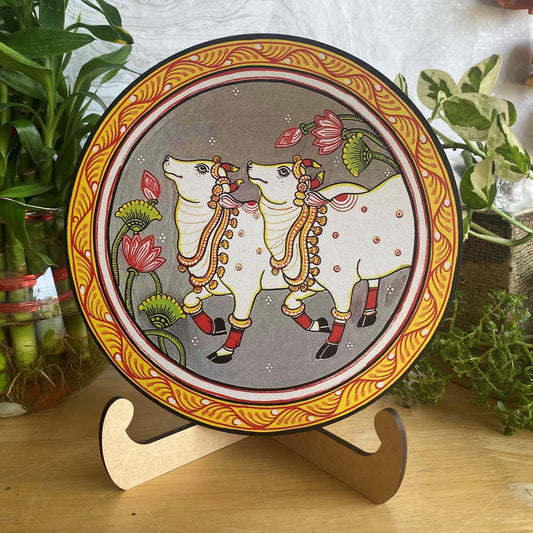 Hand-painted decorative plate with two white cows on a grey background, surrounded by lotus flowers and an ornamental border, displayed on a wooden stand.