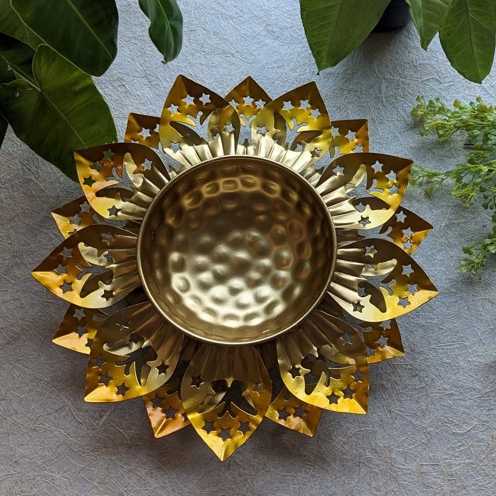 Handcrafted floral metal urli with pink petals and a golden hammered center bowl, placed among green plants.
