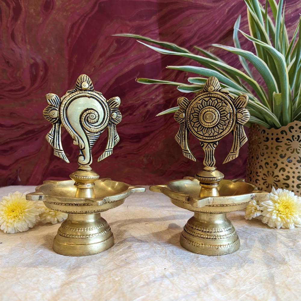 Set of two handcrafted brass oil lamps featuring Conch and Chakra designs with intricate carvings, placed on a decorative background.