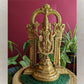 Intricately designed brass Maha Vishnu idol with a detailed Prabhavali arch, displayed on a woven mat with green foliage and a red background.