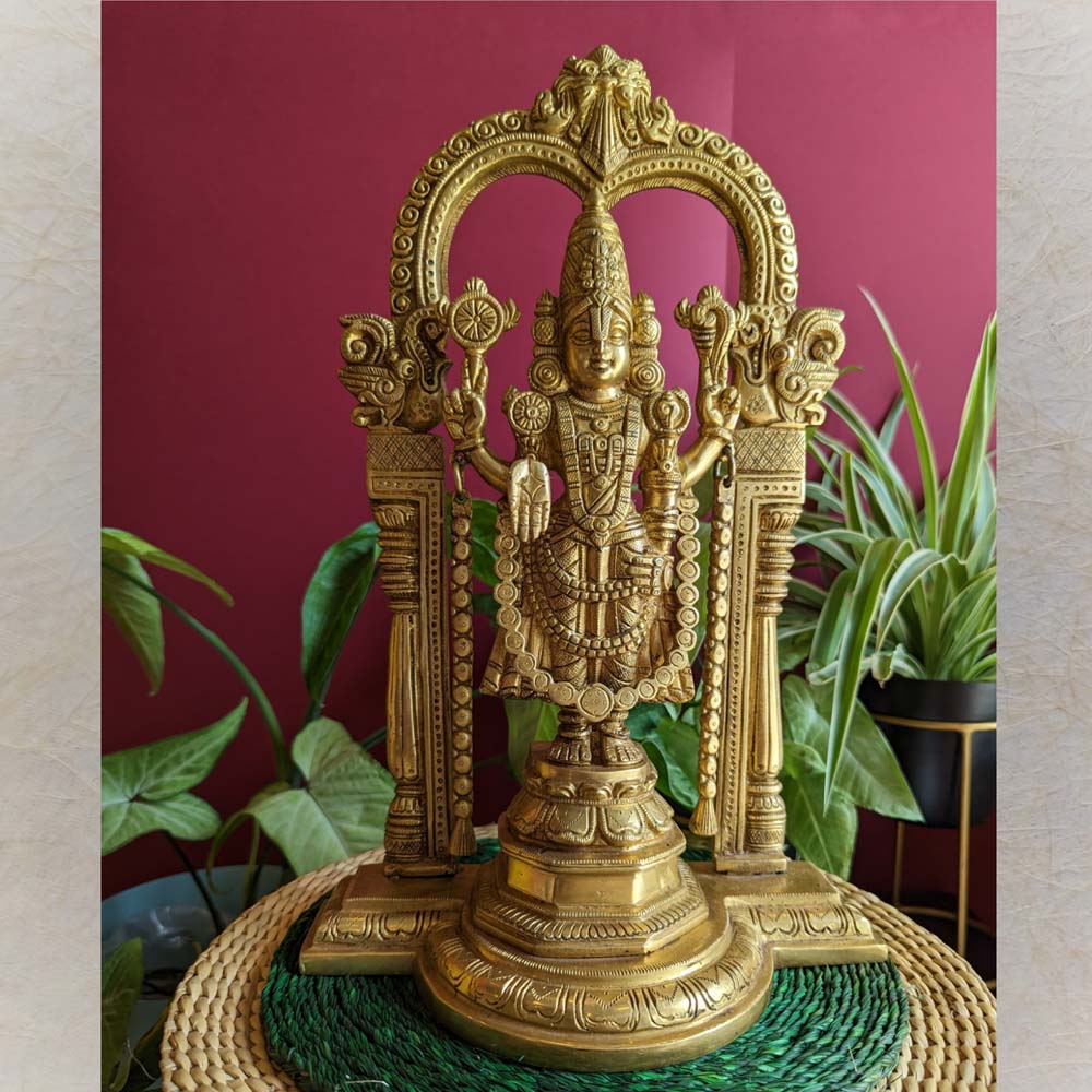Intricately designed brass Maha Vishnu idol with a detailed Prabhavali arch, displayed on a woven mat with green foliage and a red background.