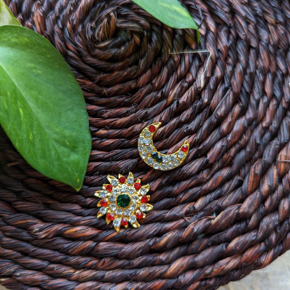 Gold-toned Sun and Moon decorative pieces adorned with crystal rhinestones and ruby accents, displayed on a woven brown mat with green leaves.