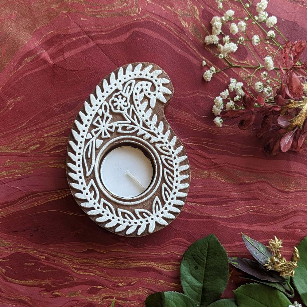 Handcrafted paisley-shaped wooden tea light holder with intricate white designs, displayed on a rich red background with floral accents.