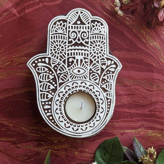 Handcrafted Hamsa-shaped wooden tea light holder with intricate white designs, displayed on a red textured background with floral accents.