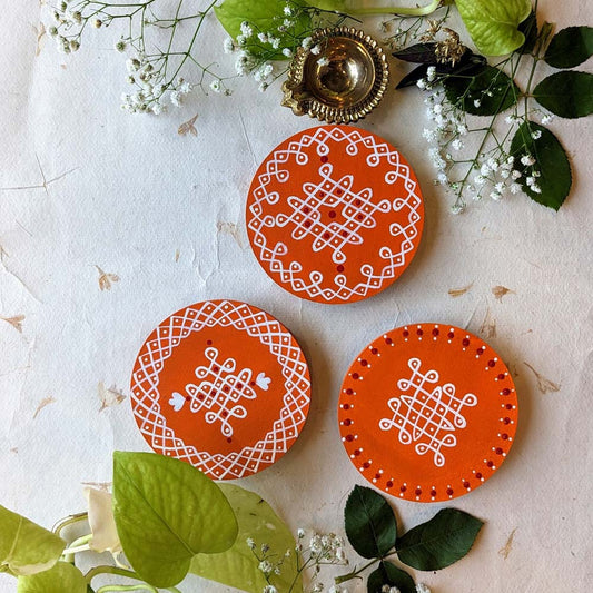 Handcrafted orange round wooden coasters featuring central kolam designs in white, set on a natural decorative background with floral accents.