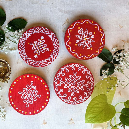 Handcrafted red round wooden coasters with white kolam center designs and yellow accents, displayed on a natural decorative background with floral elements.