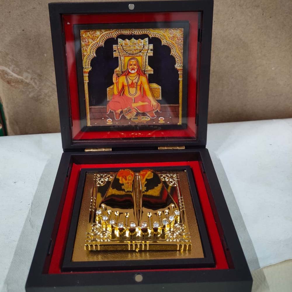 Sri Raghavendra Padhuka set in a black-framed box with a golden depiction of holy feet and a vibrant framed image of Sri Raghavendra in the background.