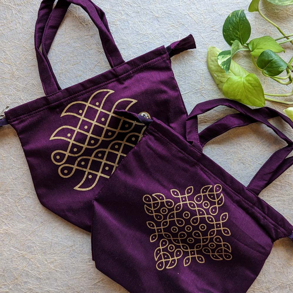Purple cotton tote bags featuring intricate golden kolam designs, displayed on a textured background with green leaves.