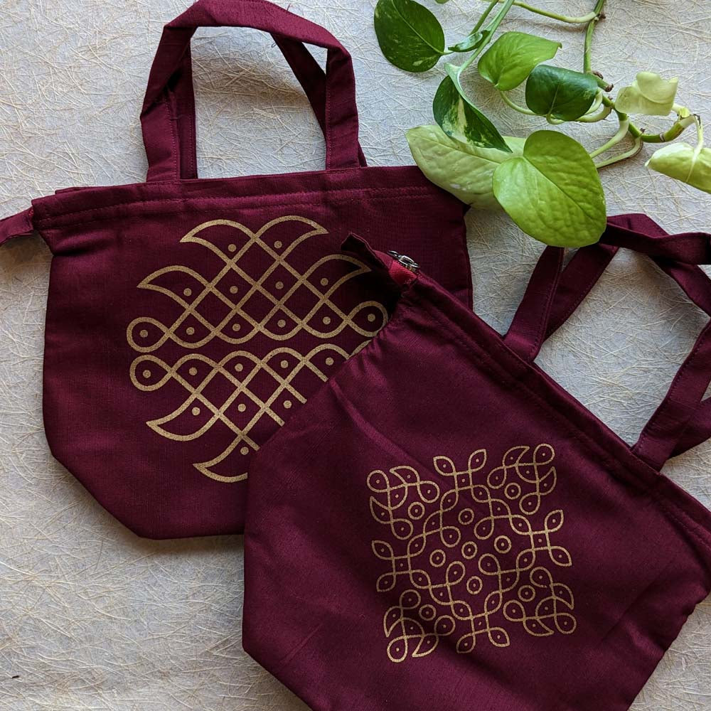 Maroon cotton tote bags featuring golden kolam designs, displayed on a textured background with green leaves.