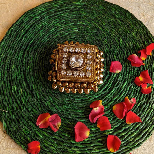 Square ghungroo kumkum box with crystal accents and gold detailing, placed on a green woven mat surrounded by rose petals.