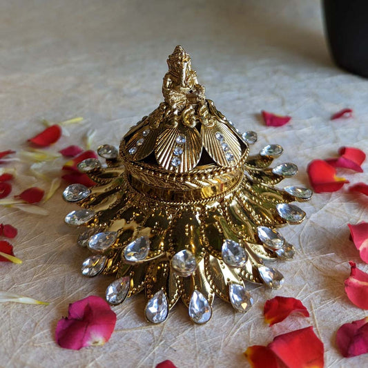 Gold Ganesh kumkum box with intricate detailing, crystal embellishments, and a Ganesha figurine, surrounded by rose petals.