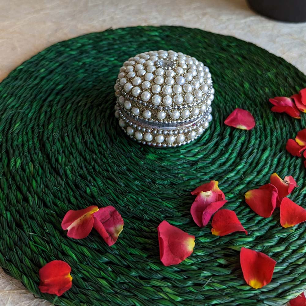 Round pearl-studded kumkum box with intricate detailing, placed on a green woven mat adorned with red rose petals.