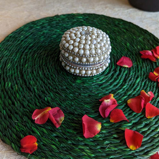 Round pearl-studded kumkum box with intricate detailing, placed on a green woven mat adorned with red rose petals.